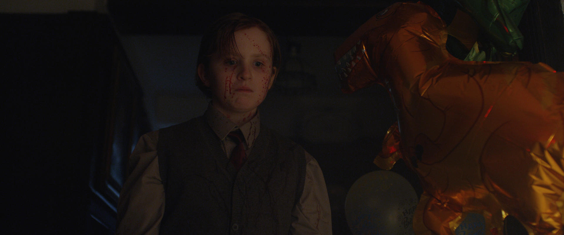 Ein Junge mit blutigen Wangen steht neben einem orangefarbenen Drachenballon.