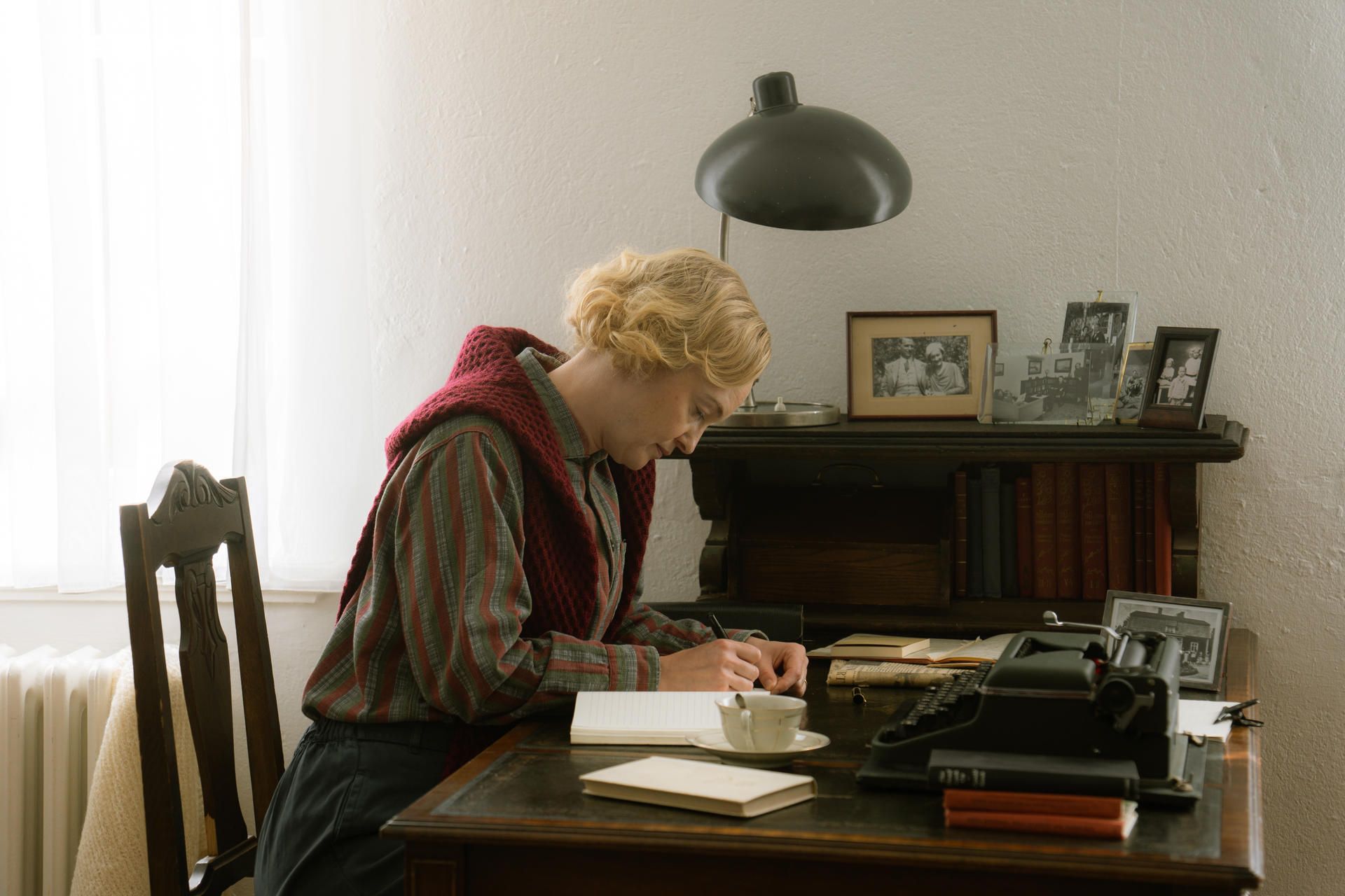Eine Frau mit blonden Haaren schreibt an einem Schreibtisch. Sie trägt einen roten Schal.