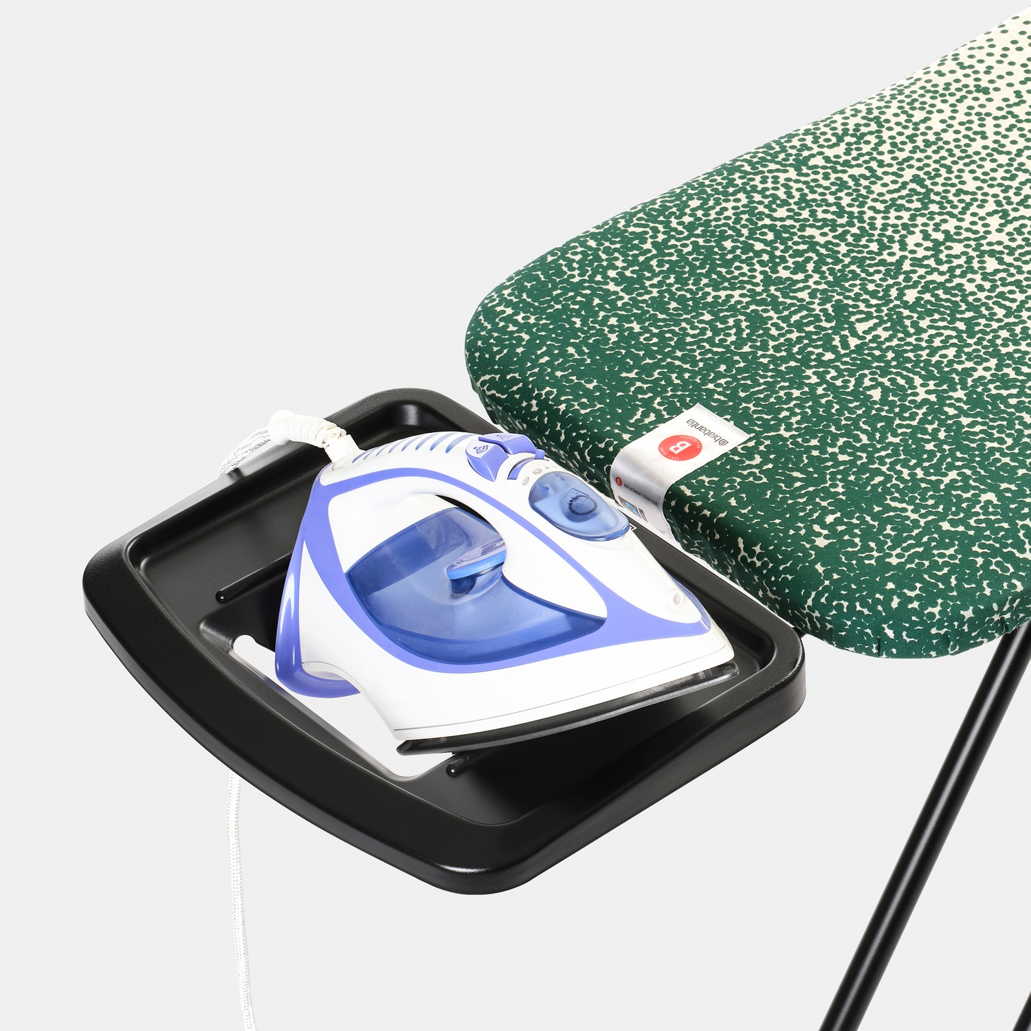 A white and blue iron sits on a black tray next to a green patterned ironing board against a white background.