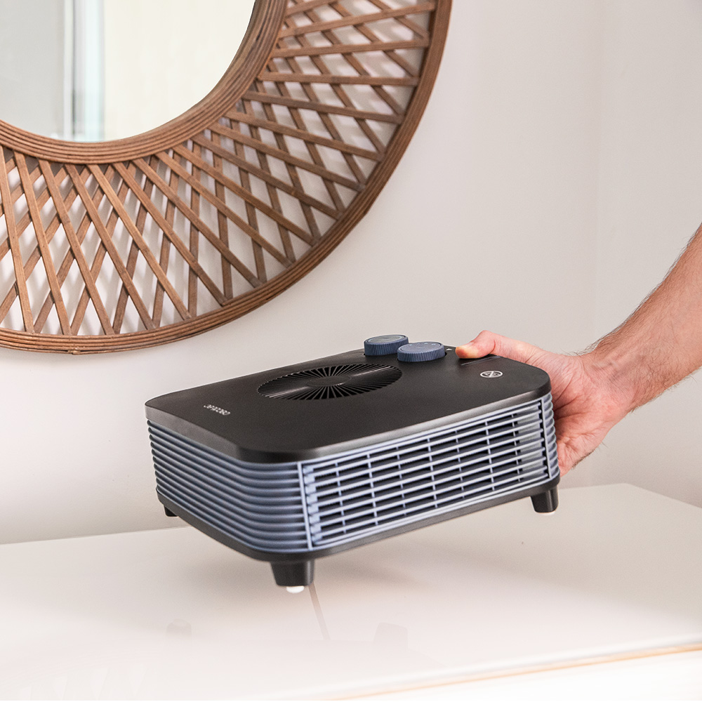 Un ventilador negro con perillas azules está sobre una mesa blanca. Un hombre lo sujeta.
