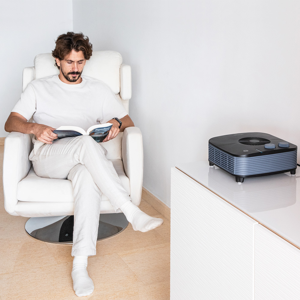 Hombre lee libro en sillón blanco, purificador de aire en mesa.