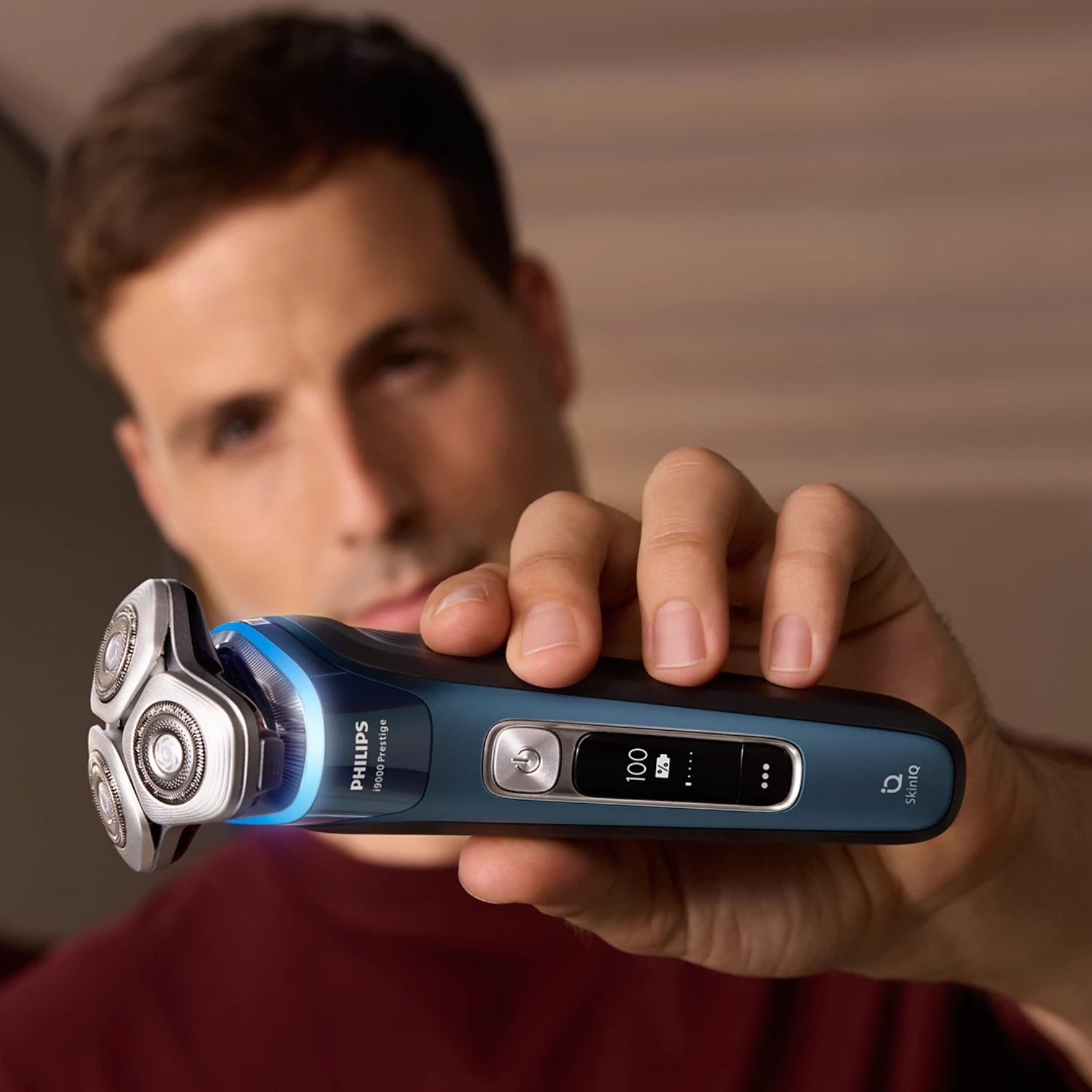Hombre sostiene una afeitadora Philips azul con cabezal plateado. Pantalla muestra 100%.