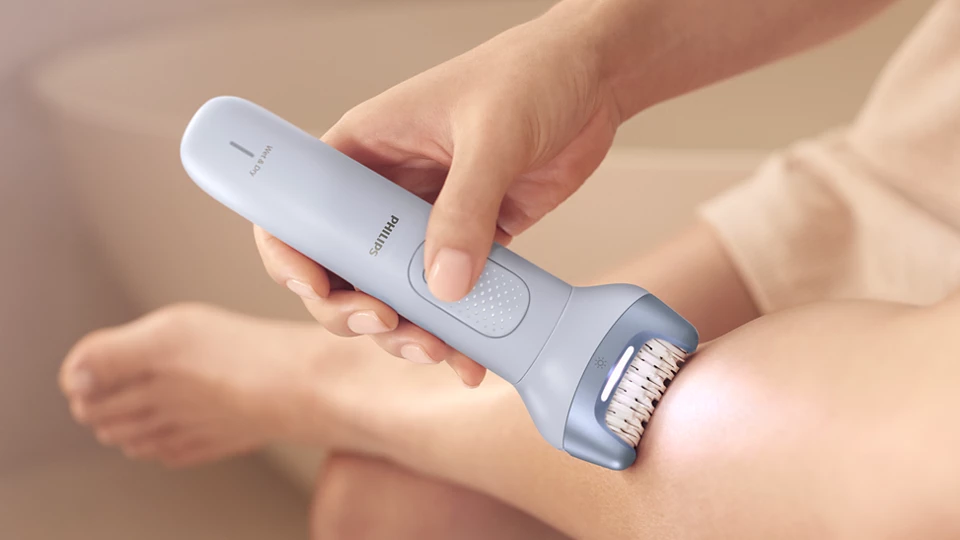 Egy világoskék Philips epilátor használatban van egy lábon. A készüléknek fehér gombja és fénye van.