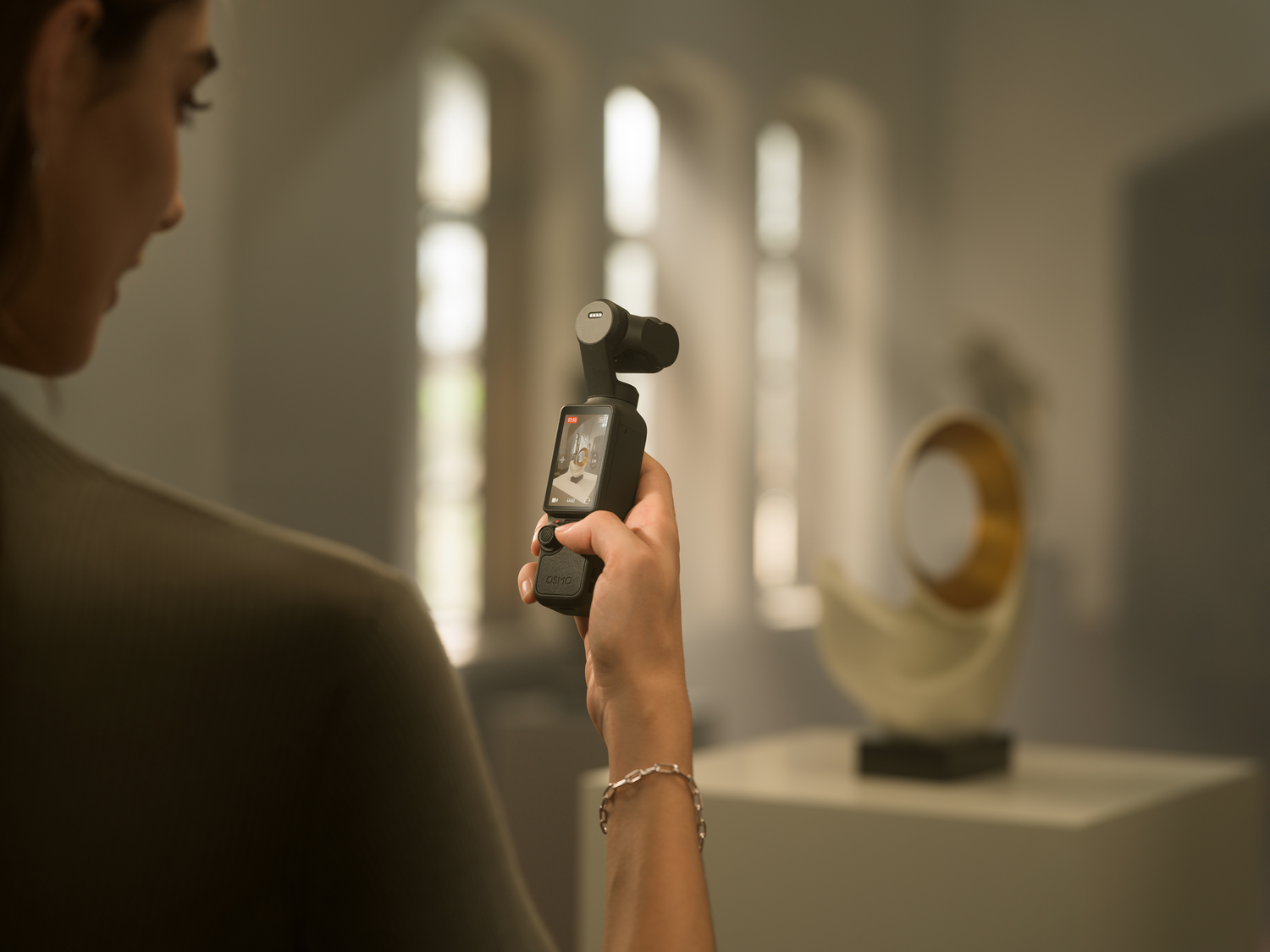 Mujer sostiene cámara filmando una escultura. Fondo gris.