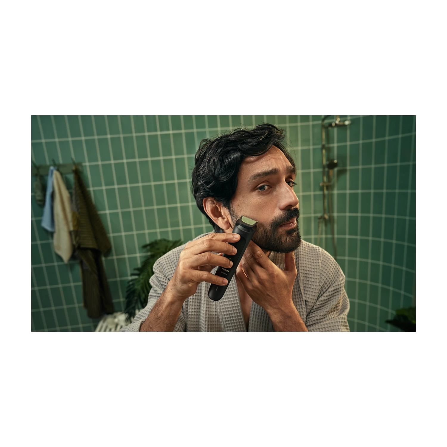 Uomo in accappatoio si rade con rifinitore in bagno con piastrelle verdi.