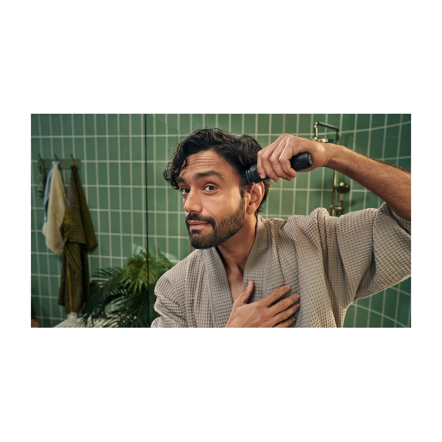 Uomo con barba in accappatoio usa un rifinitore sulla testa. Piastrelle verdi.
