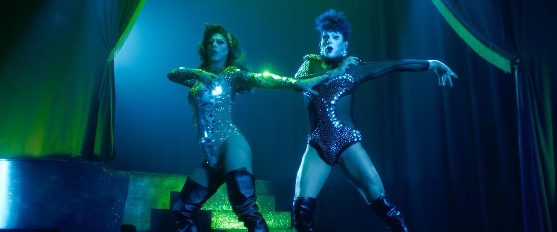 Zwei Dragqueens in glitzernden Kostümen tanzen auf einer Bühne im Scheinwerferlicht.
