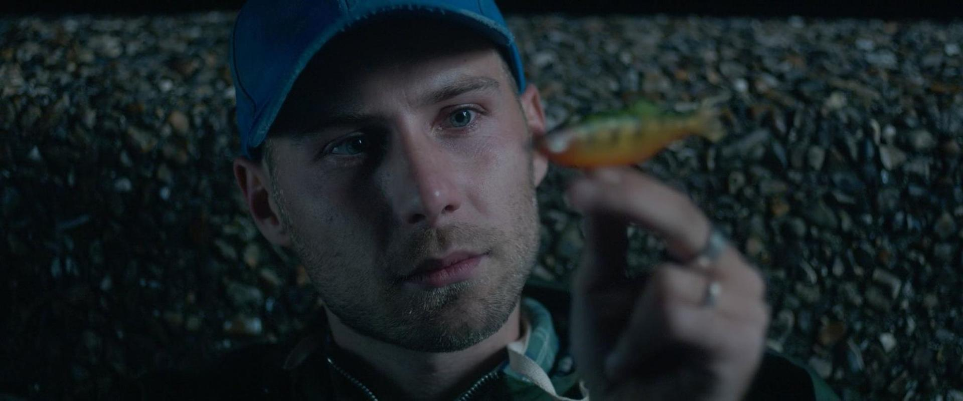 Ein Mann mit blauer Kappe hält einen kleinen, bunten Fischköder in seiner Hand.