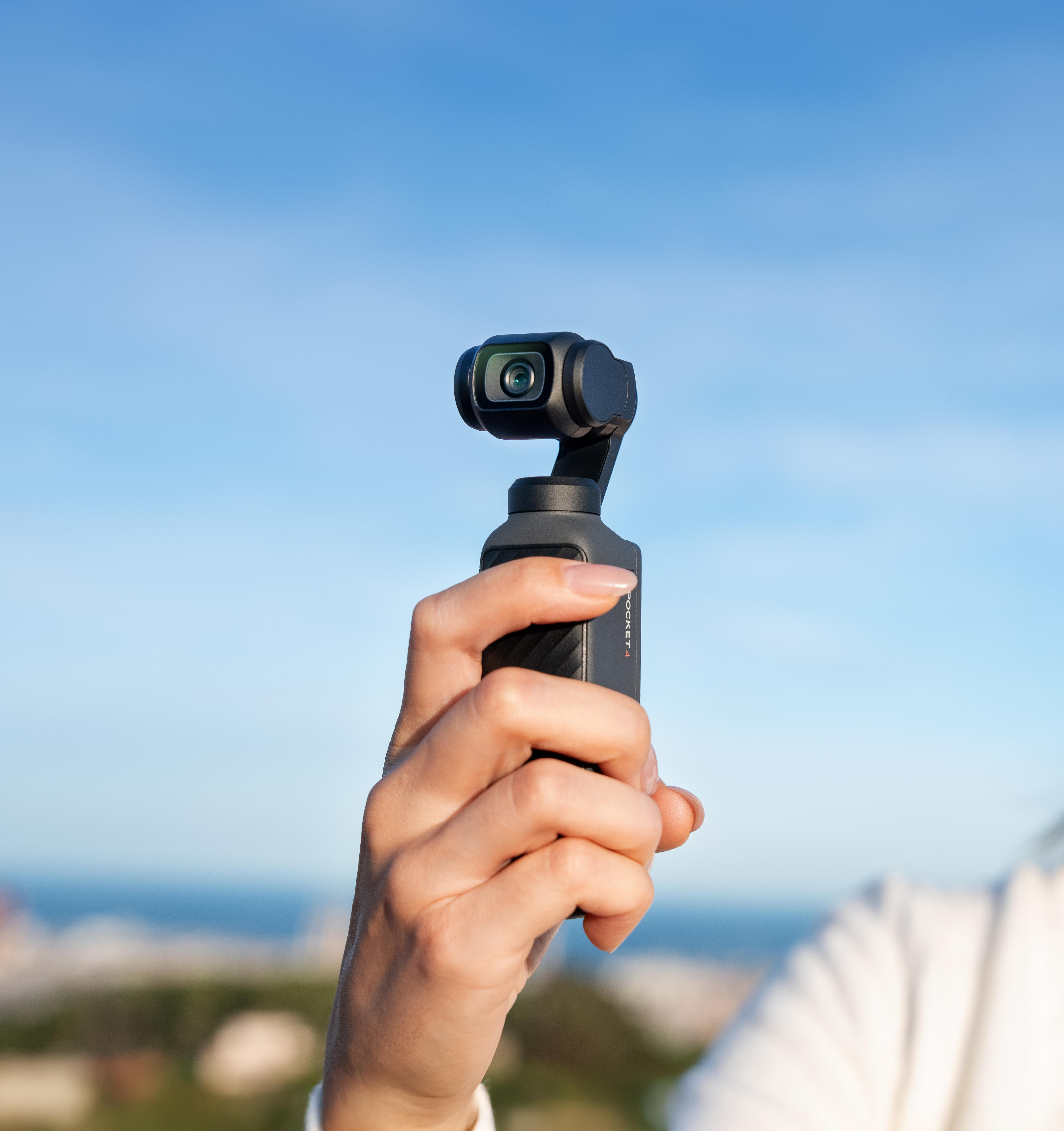 Eine Hand hält eine schwarze Kamera mit Gimbal vor blauem Himmel.