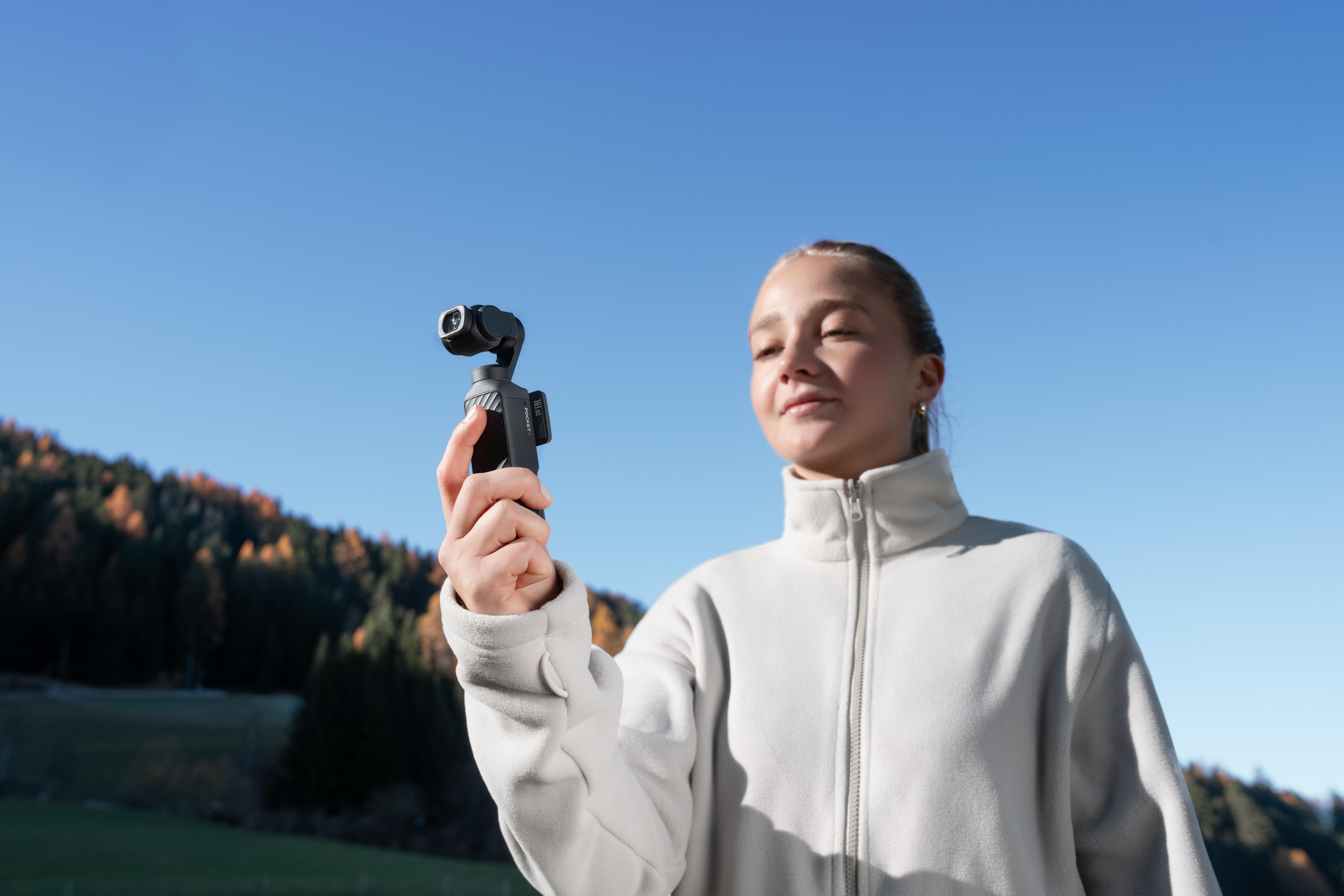 Eine Frau hält eine Kamera mit Gimbal vor klarem Himmel und bewaldeten Hügeln.
