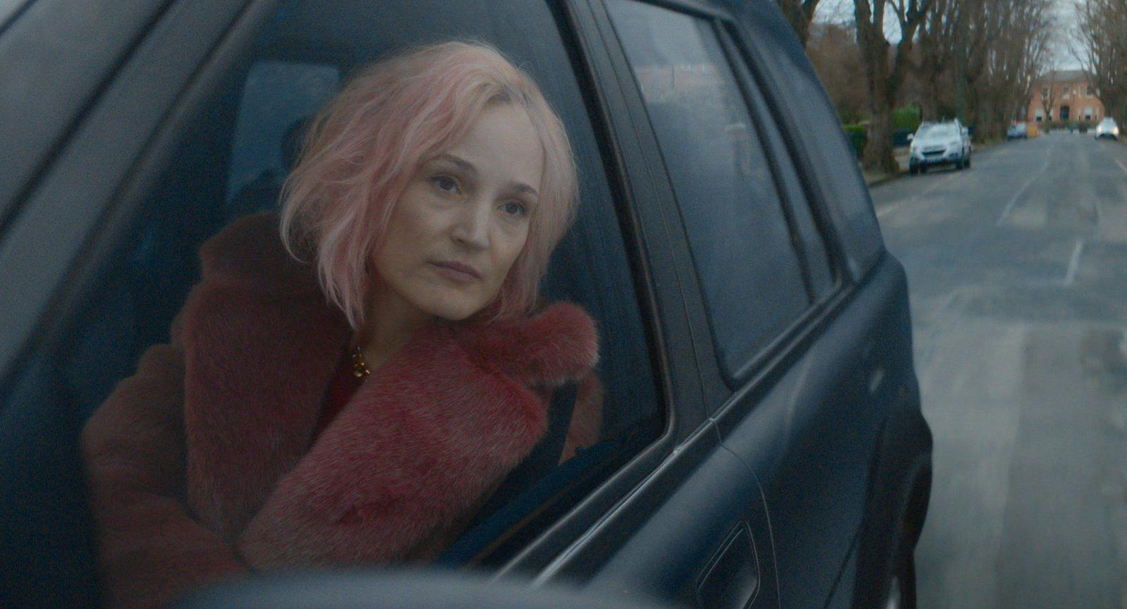 Eine Frau mit rosa Haaren sitzt im Auto und trägt einen roten Pelzmantel.
