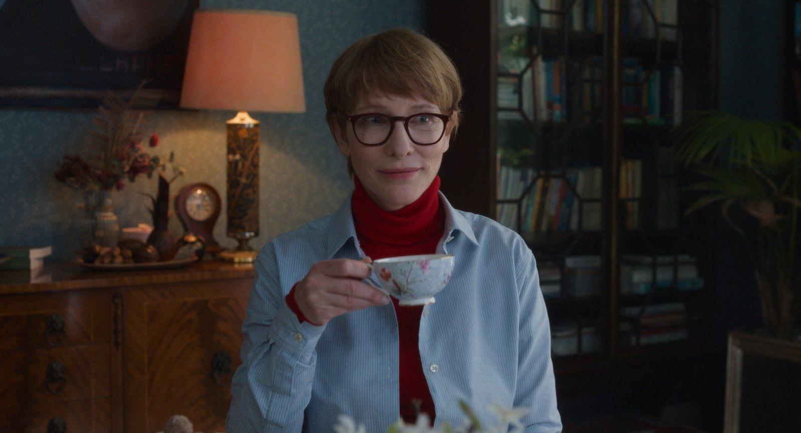 Eine Frau mit Brille trinkt aus einer kleinen Tasse in einem Zimmer mit Bücherregal.