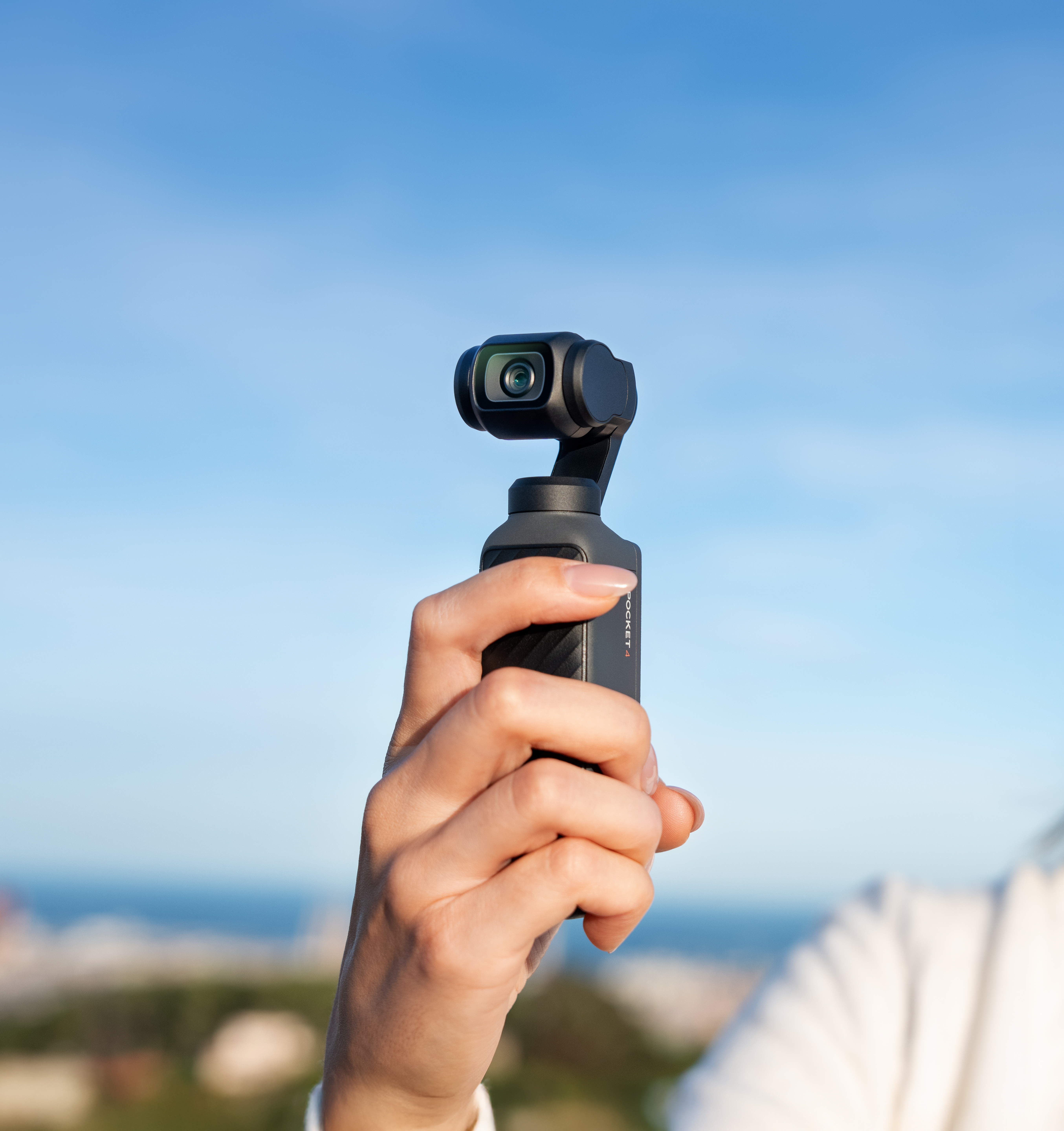 Eine Hand hält eine kleine schwarze Kamera mit Gimbal vor blauem Himmel.