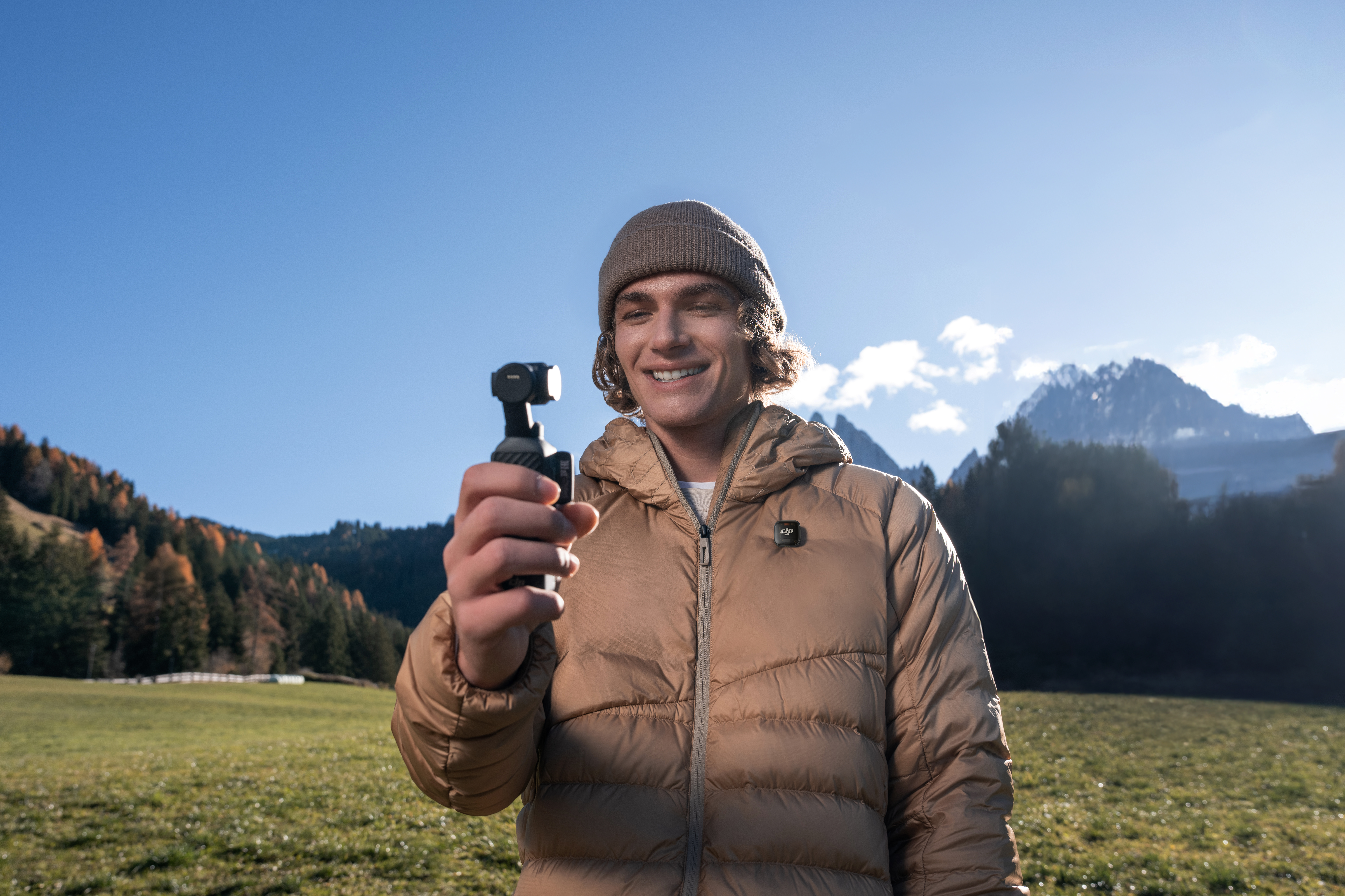 Ein Mann mit Mütze und Daunenjacke hält eine Kamera in einer Berglandschaft.
