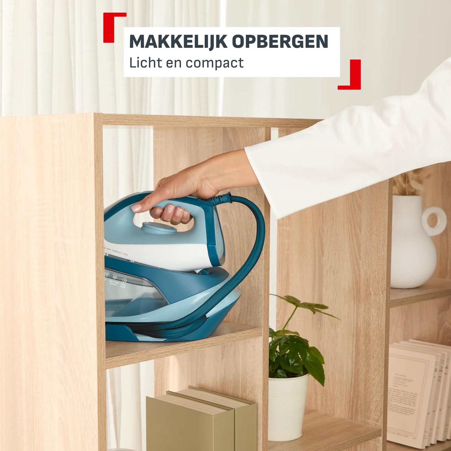 Een hand plaatst een blauw stoomstrijkijzer op een plank. Naast een plant in pot.