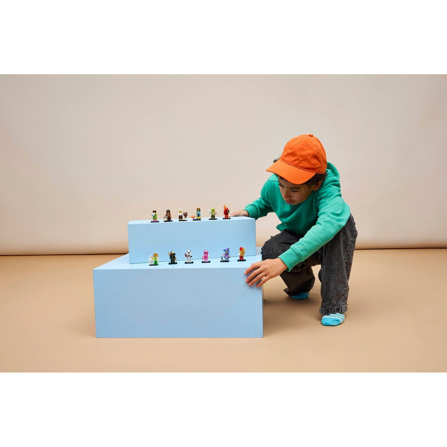 Un ragazzo con cappello arancione e felpa verde si inginocchia davanti a gradini blu con figure giocattolo.