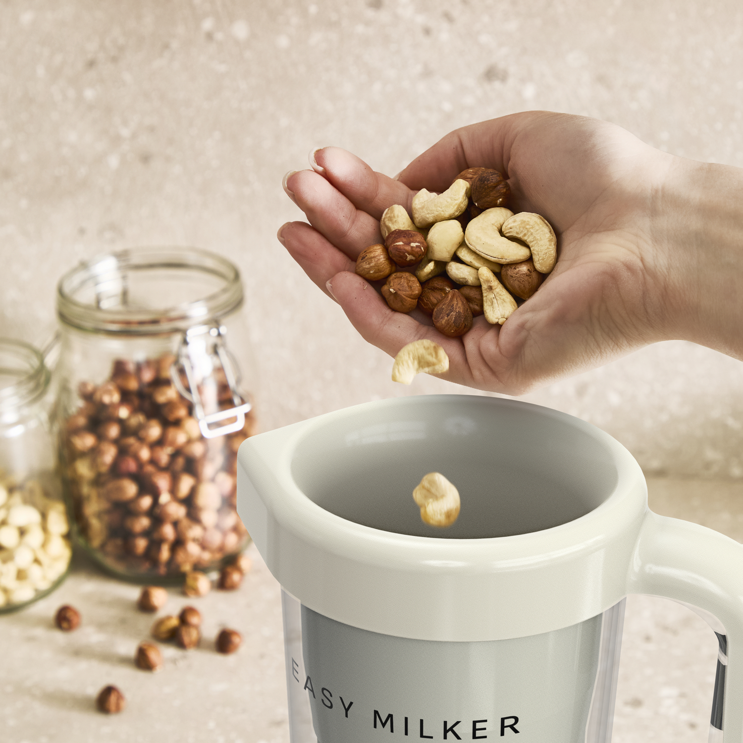 Een hand houdt hazelnoten en cashewnoten boven een melkmachine. Potten met noten op de achtergrond.