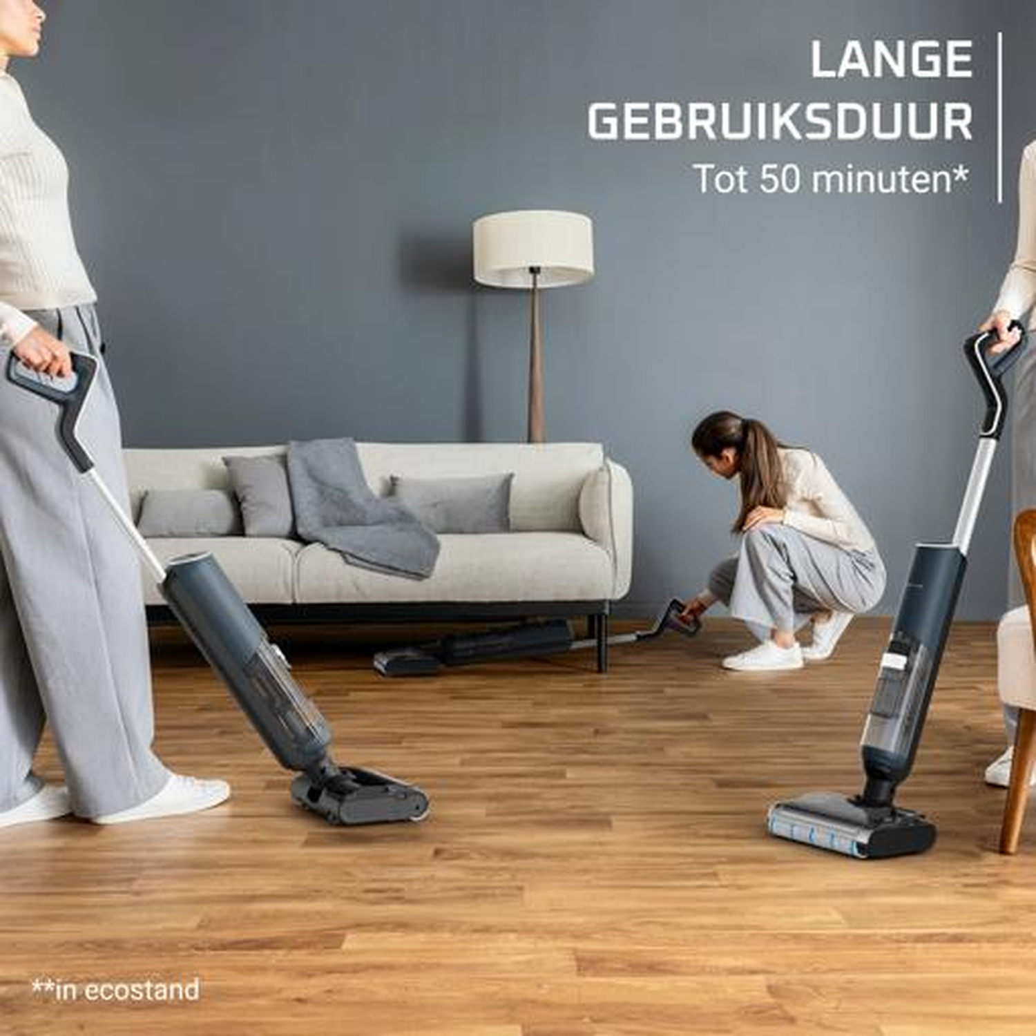 Two people use vacuum cleaners in a living room with a wooden floor.