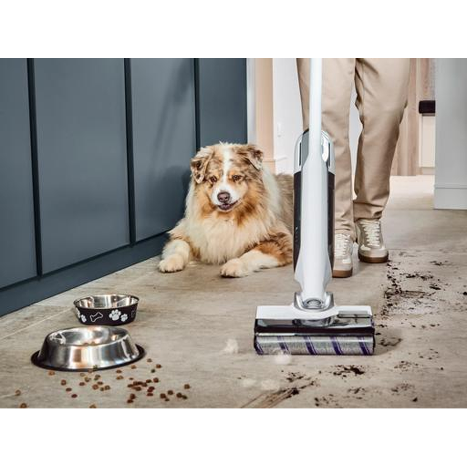 A dog lies near food bowls while a person vacuums.