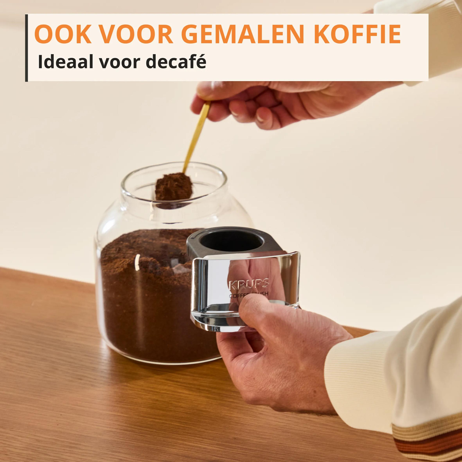 Een hand schept koffiepoeder uit een glazen pot met een lepel.