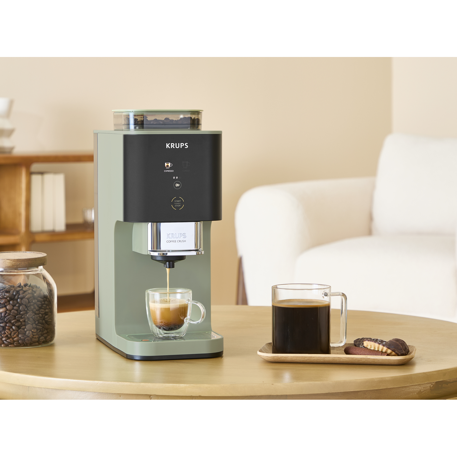 Een groene koffiezetapparaat zet espresso in een kopje. Daarnaast een kop koffie en koekjes.