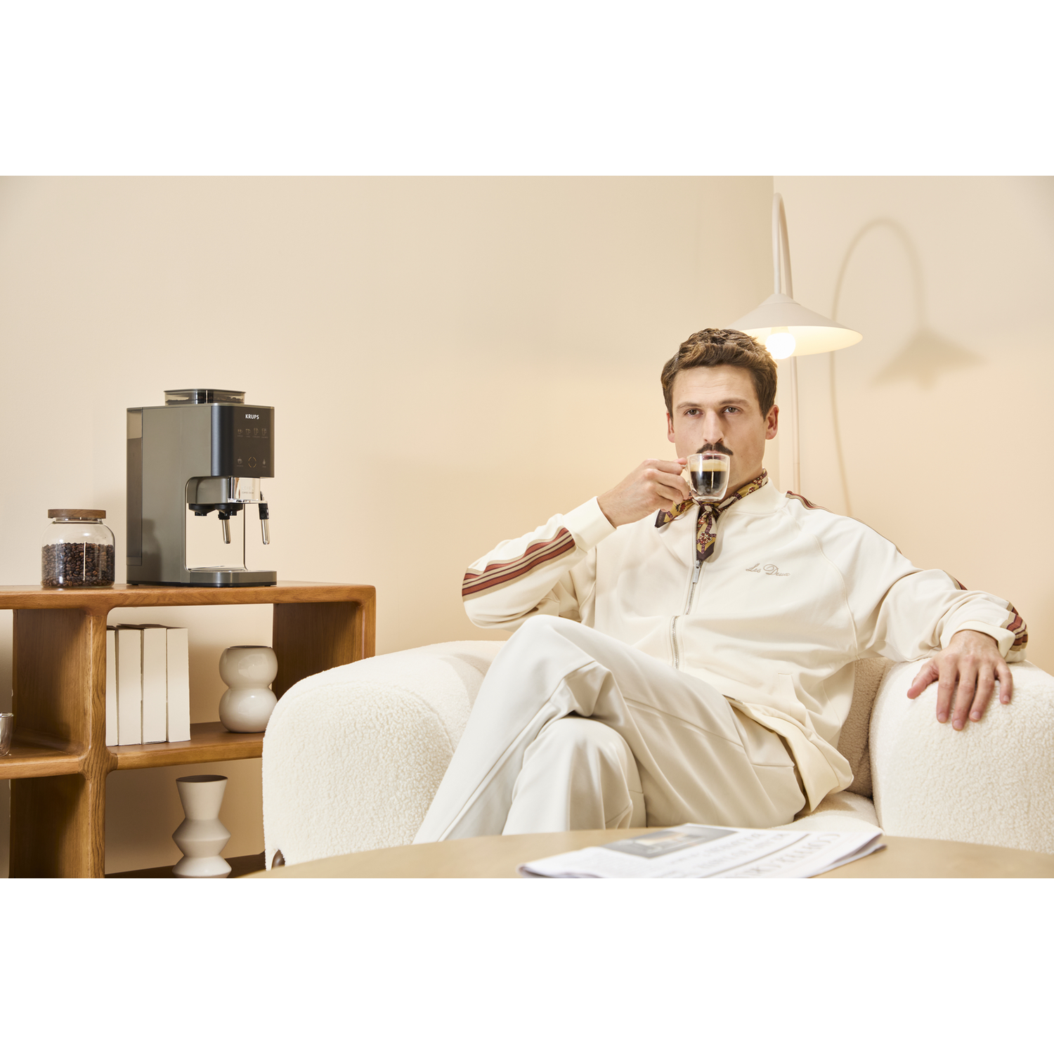Man in white tracksuit drinks espresso next to coffee machine and shelf.