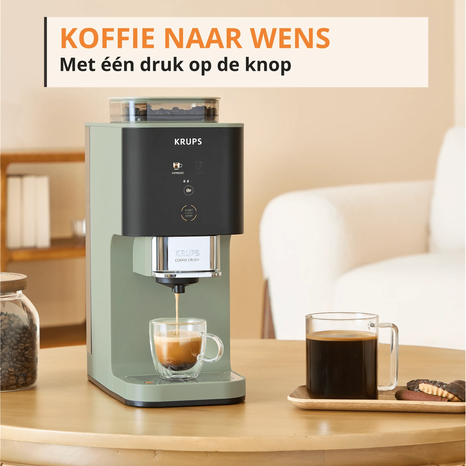 A green Krups coffee machine brews espresso into a cup. Coffee and cookies nearby.