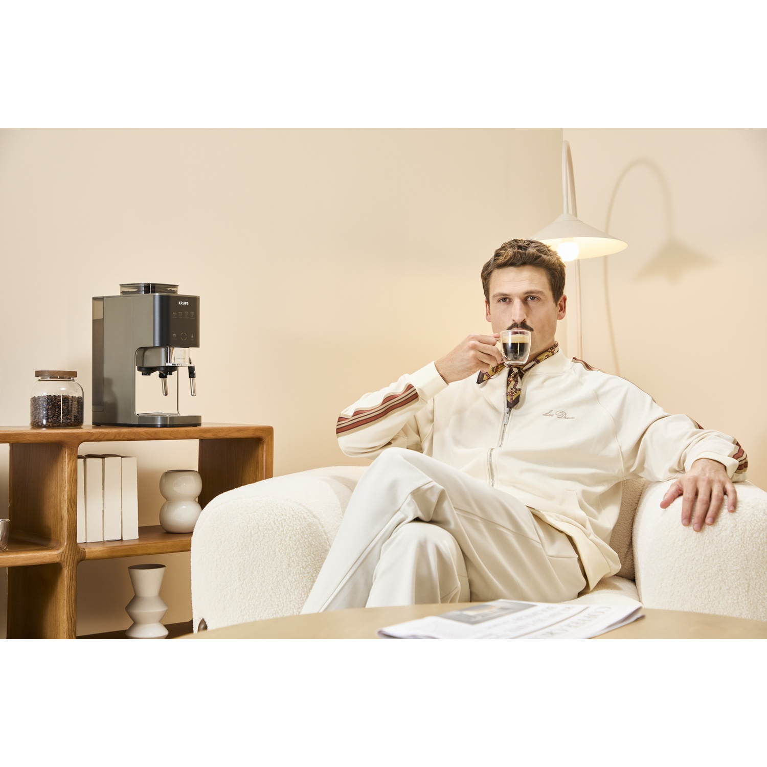 Man with mustache drinks espresso next to coffee machine on shelf with books.