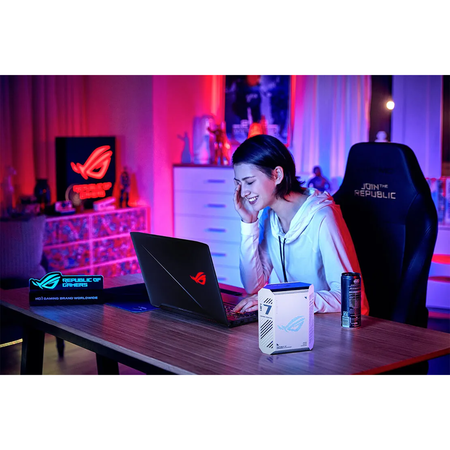 A young woman smiles while using a laptop in a room with neon lighting. A gaming PC and can sit on the desk.