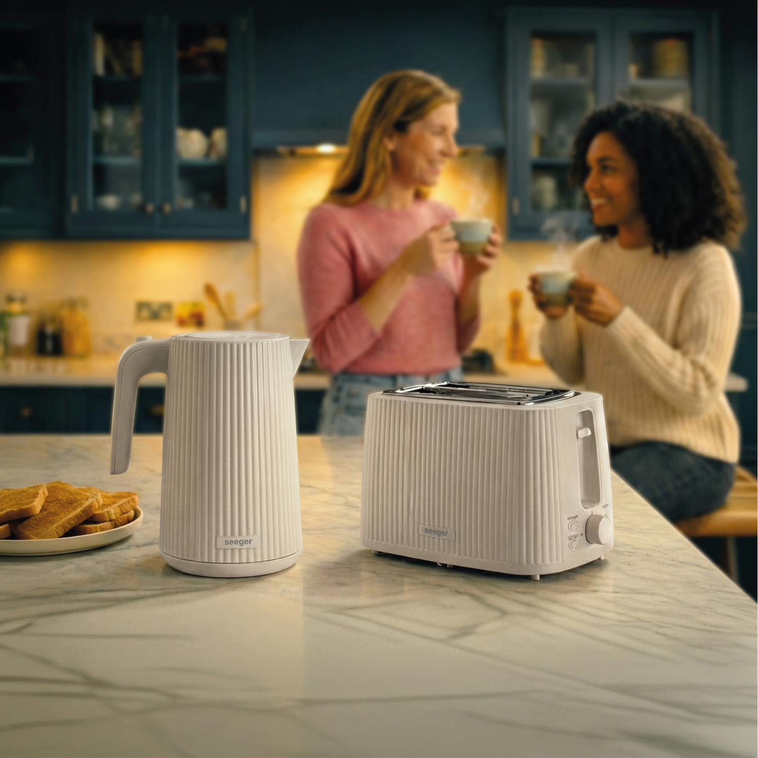 A white kettle and toaster sit on a marble counter, two women chat in the background.