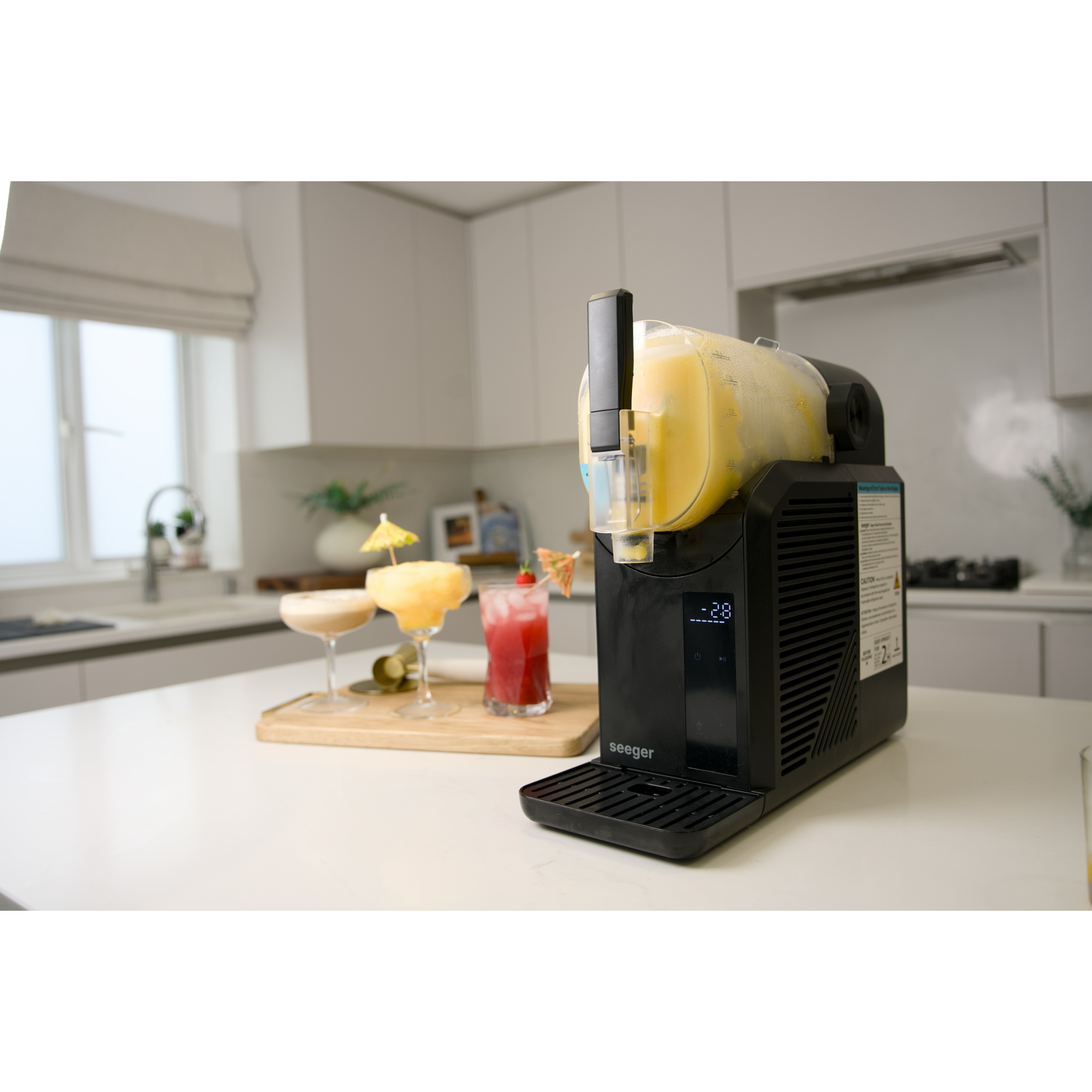 A black slush machine with frozen drink sits on a counter. Three cocktails are in the background.