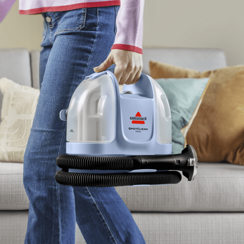 Una persona con jeans sostiene un limpiador portátil Bissell SpotClean Mini azul claro.