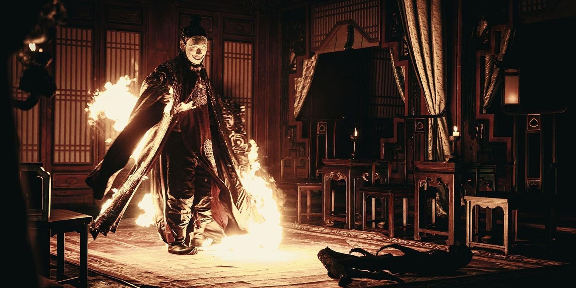 Maskierte Figur in Flammen inmitten eines opulenten Raumes mit Möbeln.