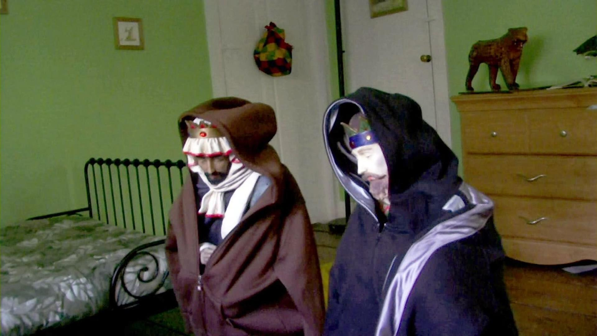 Zwei Figuren in Kapuzenmänteln mit Kronen stehen vor einem Bett und einer Kommode.