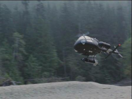 Ein dunkler Hubschrauber fliegt über einem felsigen Ufer vor einem Wald.