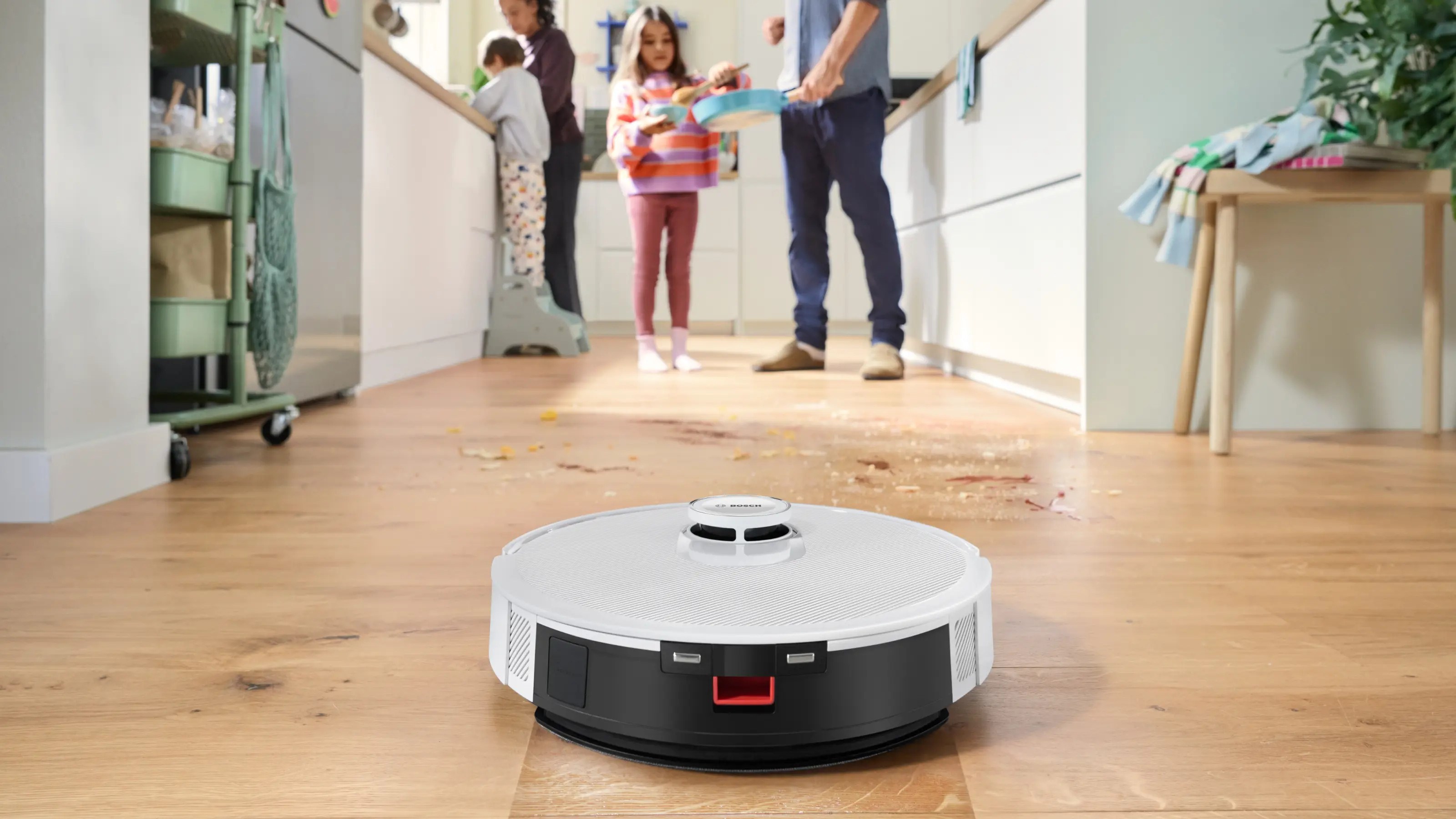 Robot sprzątający czyści drewnianą podłogę w kuchni, podczas gdy rodzina krząta się w tle.