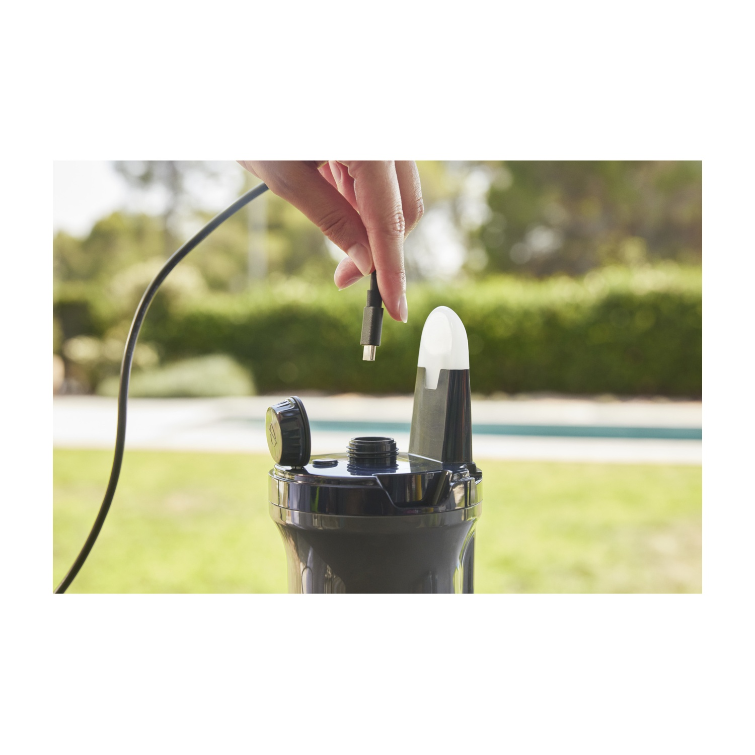 Mano che tiene cavo USB-C vicino a dispositivo nero con tappo aperto, esterno con piscina.