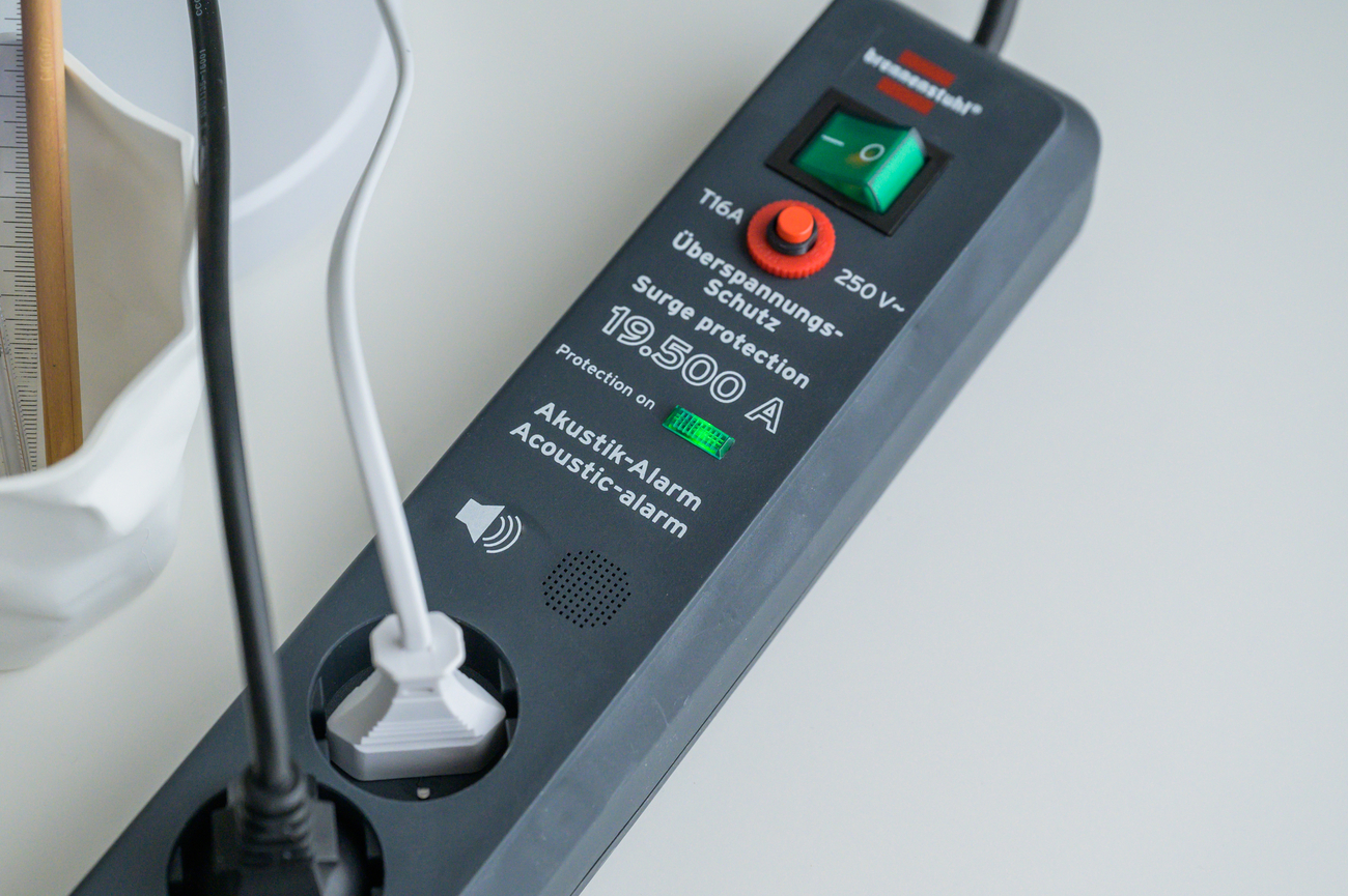 Schwarze Steckdosenleiste mit grünem Schalter, rotem Knopf, und LED-Anzeige.