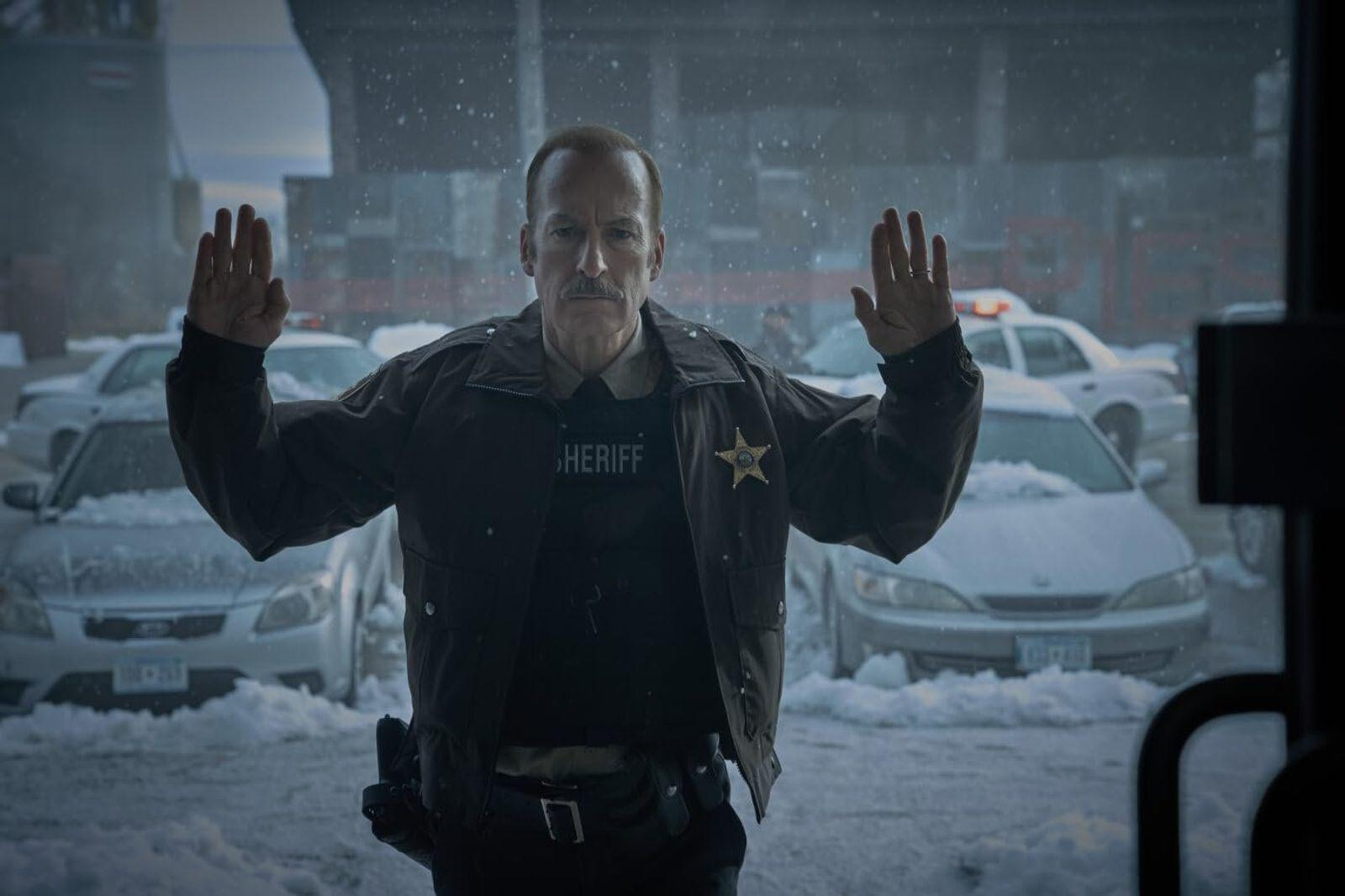 Ein Sheriff mit erhobenen Händen steht im Schnee vor Polizeiautos.