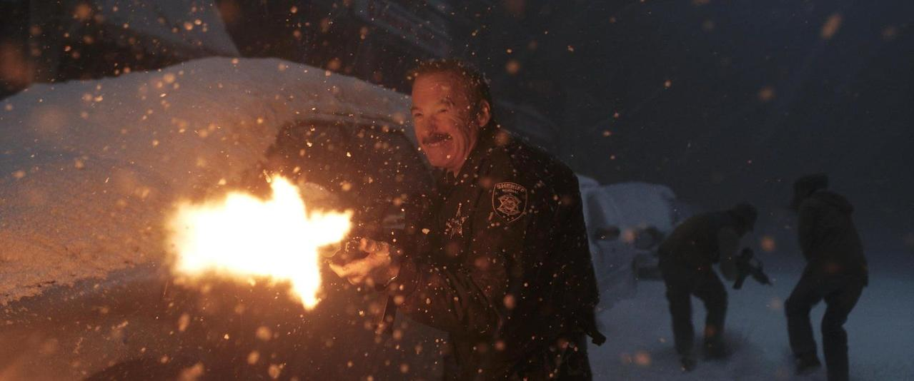 Ein Sheriff feuert in einer Schneelandschaft eine Waffe ab, während zwei Männer im Hintergrund stehen.