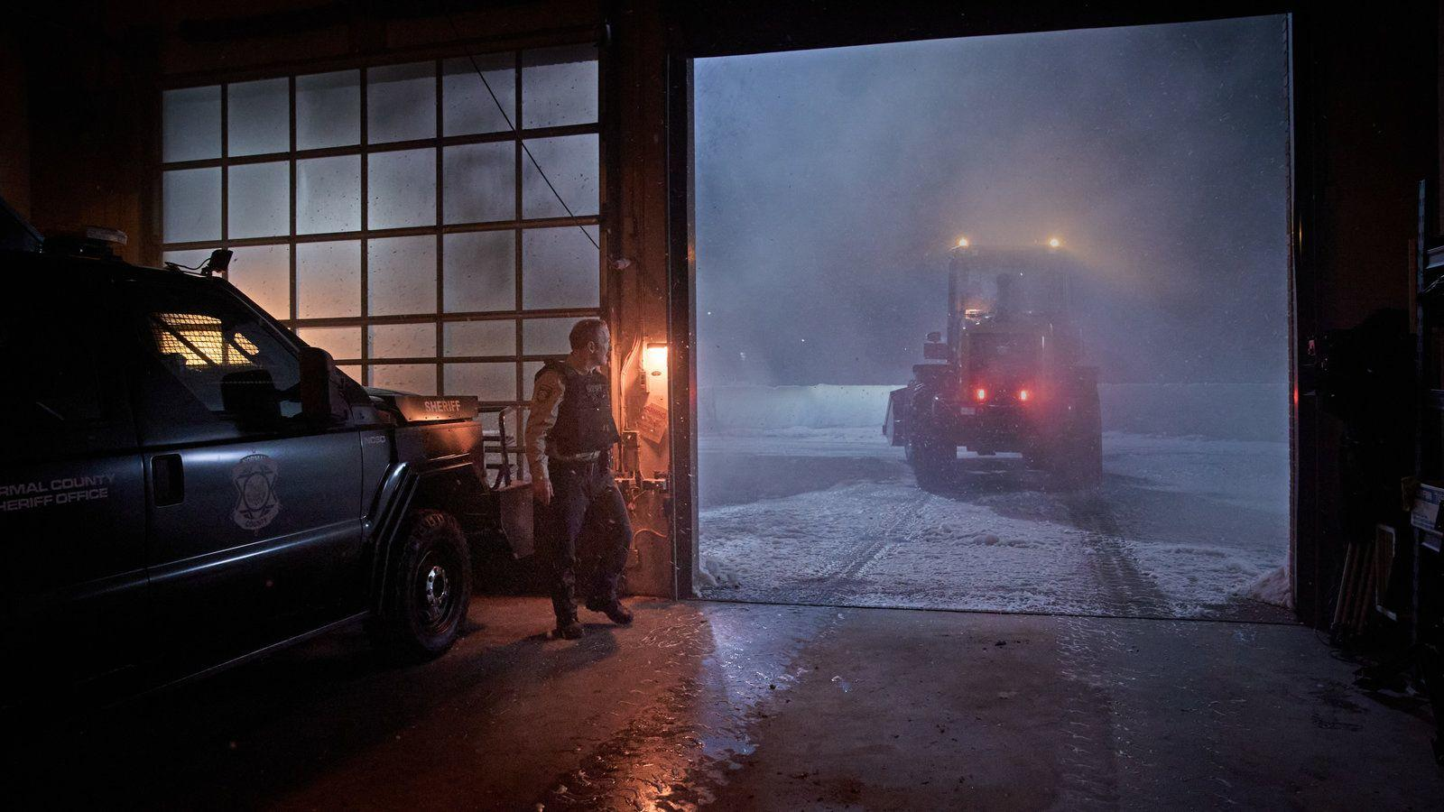 Ein Sheriff steht in einer Garagentür, während ein Traktor mit blinkenden Lichtern in schneeigen Nebel ausfährt.