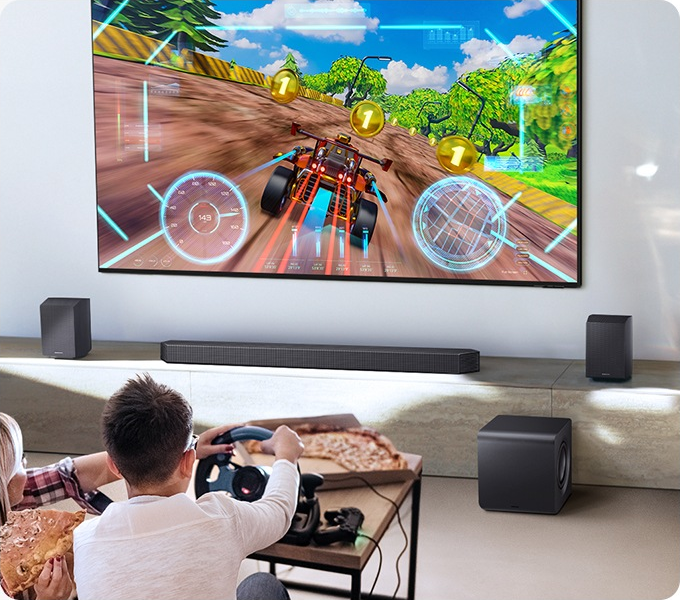 Person spielt ein Rennspiel auf einem großen Fernseher mit einem Lenkrad. Pizza liegt auf einem Tisch.