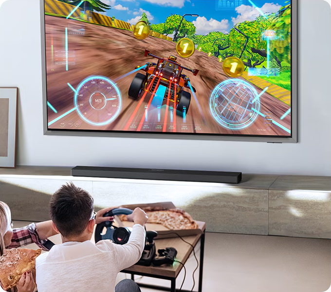 Un couple joue à un jeu de course sur TV, avec un volant et de la pizza.