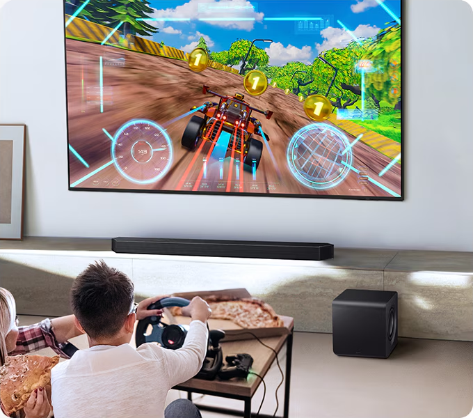 Des gens jouent à un jeu de course sur une grande télévision, avec pizza et subwoofer.
