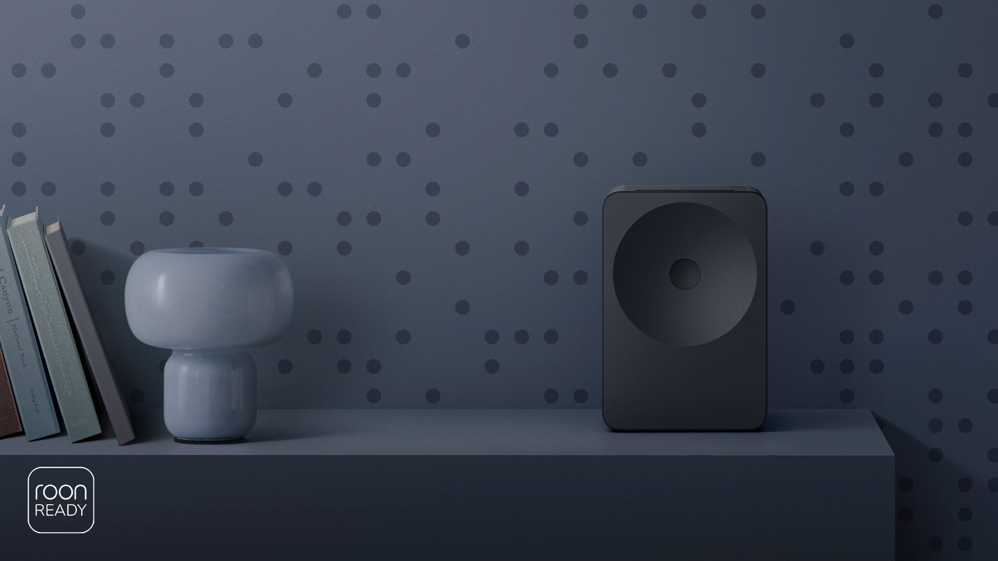 Ein dunkelgrauer Lautsprecher steht neben Büchern und einer hellblauen Lampe auf einem Regal.