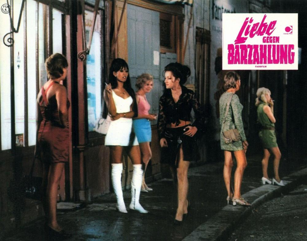 Fünf Frauen stehen auf einer Straße, einige rauchen. Ein Filmplakat ist sichtbar.