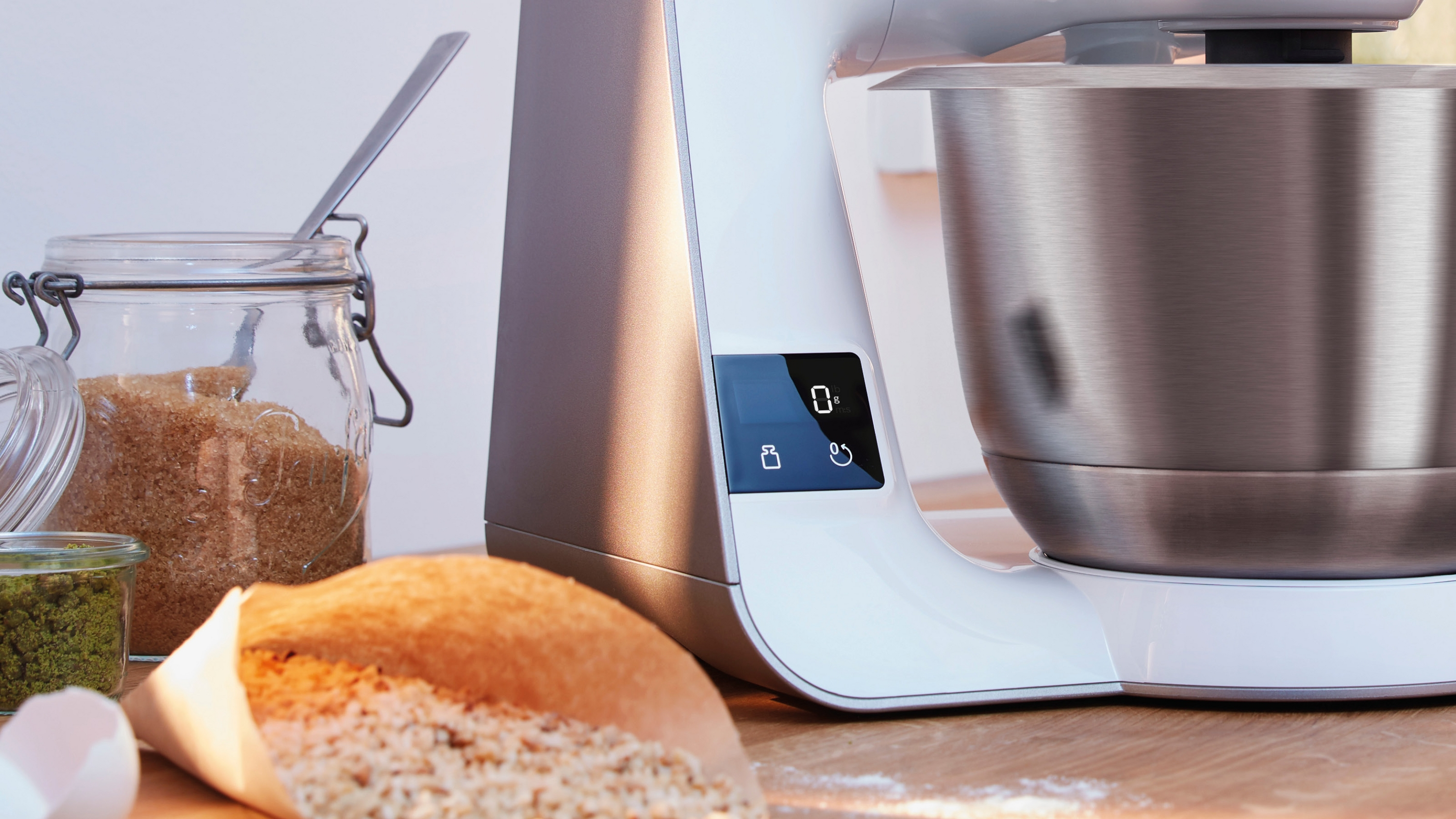 Küchenmaschine mit Edelstahlschüssel, braunem Zucker und Eiern auf Holztisch.