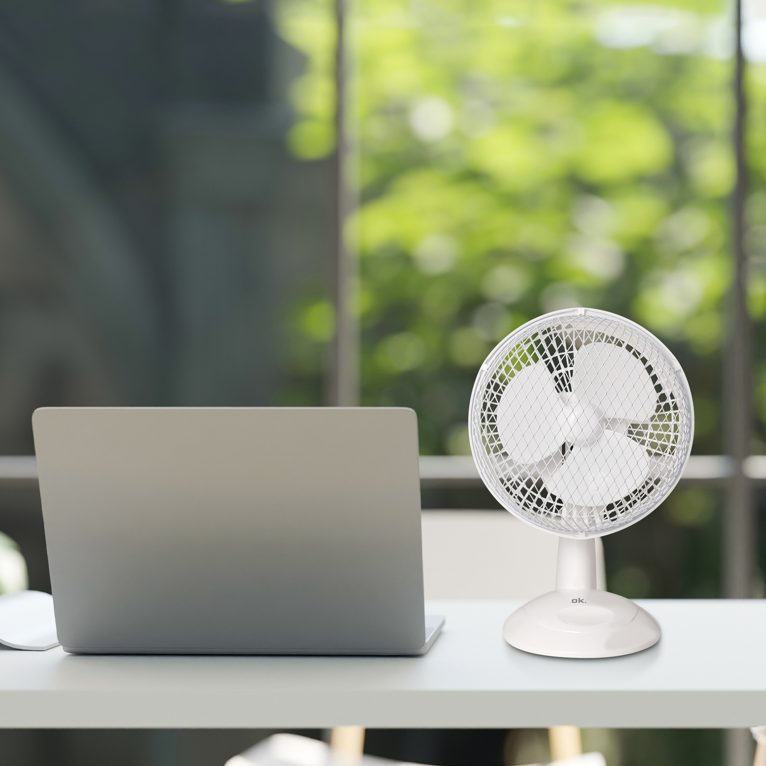 A white desk fan and a laptop on a desk, with a blurred green background.