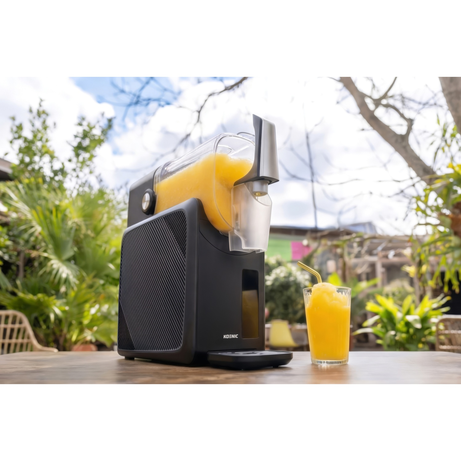 A black slush machine dispenser with a glass of orange slush next to it outdoors.