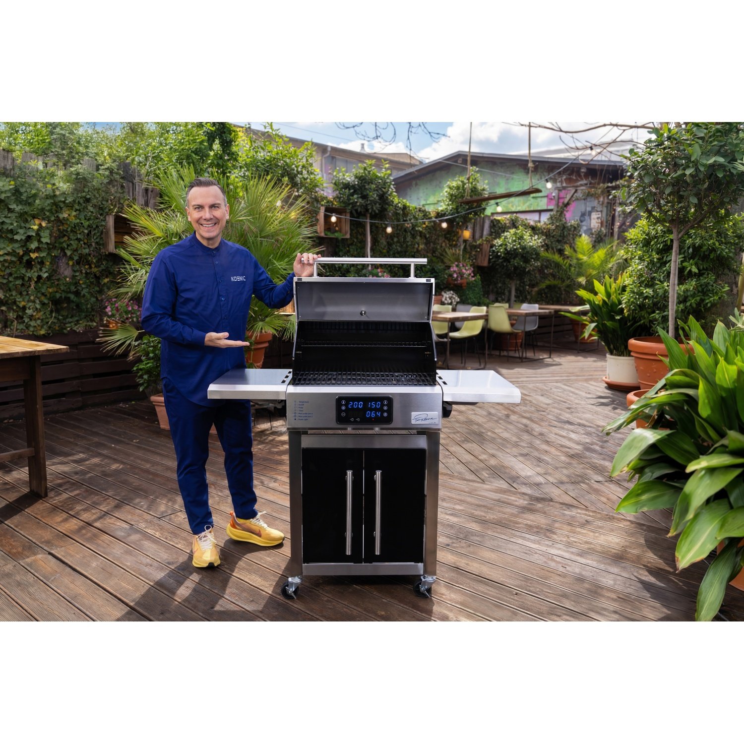 Un homme en bleu se tient à côté d'un grill sur une terrasse en bois entourée de plantes.